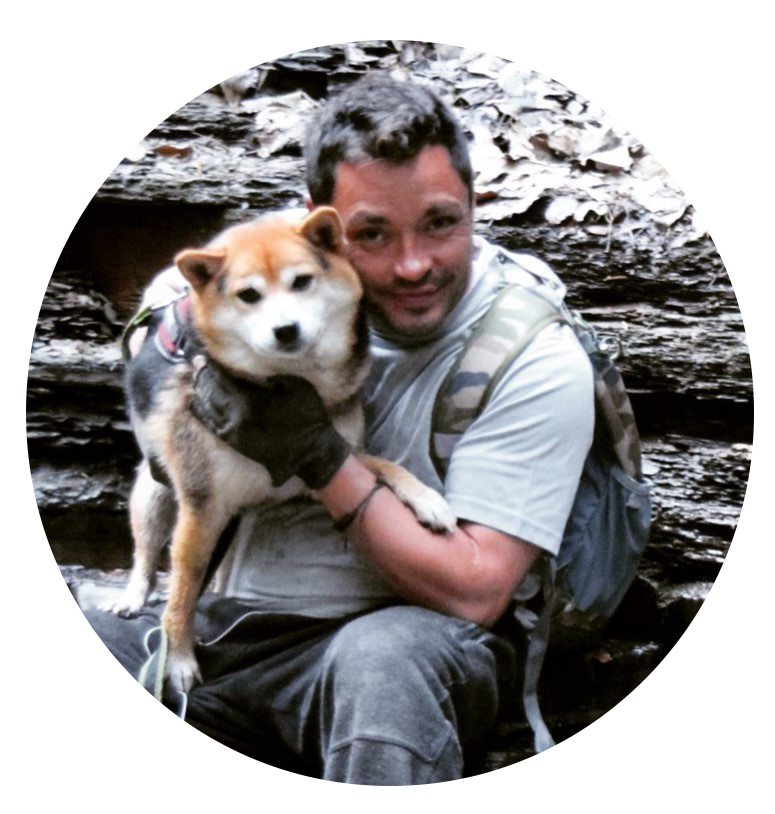 Man holding a Shiba Inu in front of logs, both wearing outdoor gear, in a forest setting.